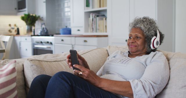 Senior woman with grey hair relaxing on couch at home, smiling while listening to music on headphones. Ideal for content related to senior living, technology use among elderly, relaxation, and leisure activities at home.