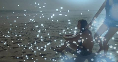 Couple Sitting on Sandy Beach Reaching Toward Water with Bokeh Light and Serene Horizon