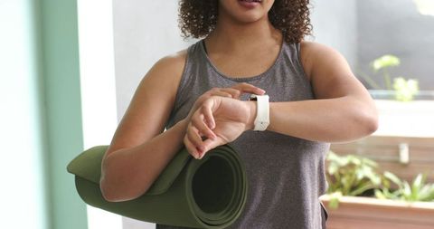 African american woman preparing for yoga class while checking smartwatch and holding mat