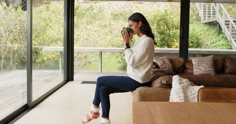 Woman sipping coffee by floor-to-ceiling glass doors in cozy minimal living space