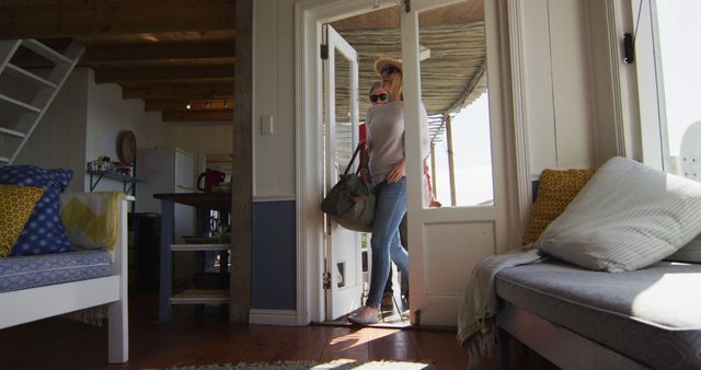 Woman entering a cozy beach house carrying a bag and wearing a sun hat. Sunny light coming through windows enhances the bright, welcoming ambiance of the interior. Ideal for promoting vacation rentals, home decor inspiration, or lifestyle blogs focusing on travel and relaxation.