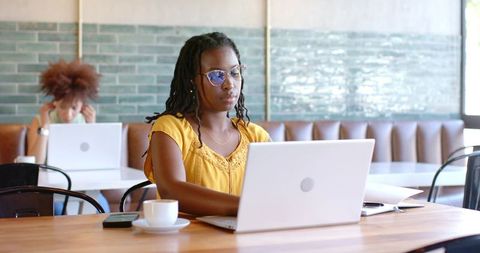 Focused black woman working on laptop in modern coffeehouse, yellow blouse, coworking