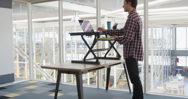 Young male professional using a standing desk while working on his laptop in a contemporary office. Transparent walls and open spaces give a modern feel to the workspace. Ideal for articles or promotional materials on workplace ergonomics, productivity in offices, or modern office design concepts.