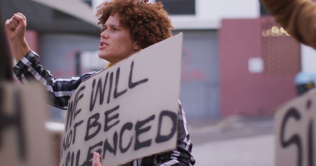 Young activist with curly hair holding placard reading 'We Will Not Be Silenced' and raising their fist passionately during urban protest. Useful for themes on social justice, activism, youth movements, and civic engagement.