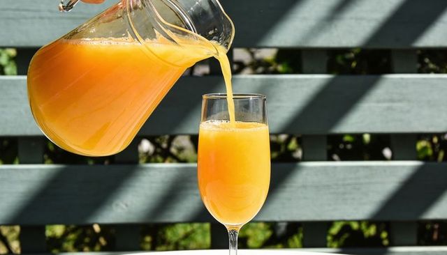 Sunlit orange juice pouring from glass pitcher into stem glass on blue-gray fence outdoor table
