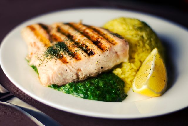 Grilled salmon filet served with yellow rice, green sauce, and lemon wedge on white plate. Ideal for use in culinary blogs, healthy eating promotion, cookbooks, recipe websites, and restaurant advertising.