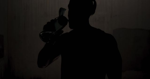 Silhouette of a man drinking from a bottle in a dark, mysterious setting. Perfect for concepts of secrecy, suspense, or introspection. Popular in storytelling, health awareness campaigns, and beverage advertisements.