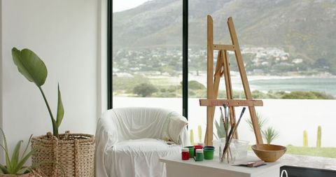 Sunlit coastal art nook showing wooden easel, paint pots, cozy white armchair and sea view