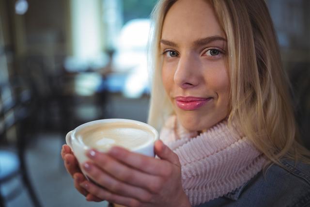 Woman in casual attire holding a hot cup of coffee in a cozy café setting. She is smiling and appears to enjoy the warm drink. Useful for images related to leisure, lifestyle, coffee culture, cafés, and relaxation.