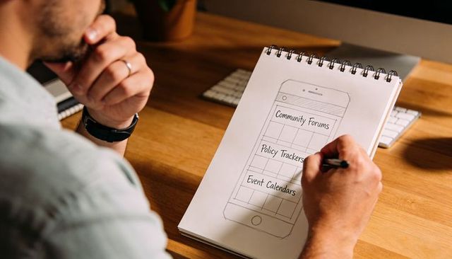 Designer sketching mobile app wireframe on spiral notepad at wooden desk with keyboard