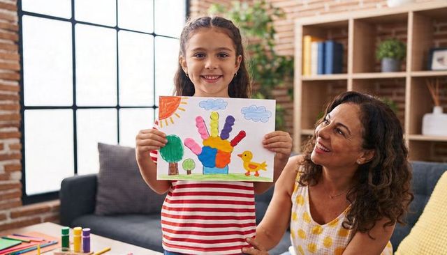 Smiling girl showing colorful handprint art while caregiver smiling in cozy living room