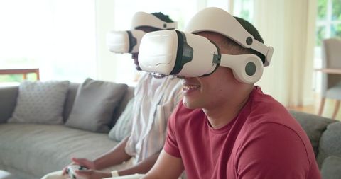 Young friends playing multiplayer virtual reality games on living room sofa with white VR headsets