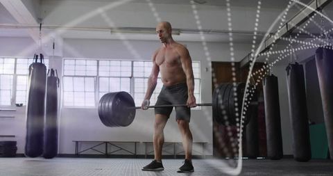 Shirtless man deadlifting heavy barbell in industrial gym with punching bags and backlight