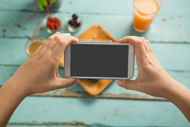 Hands holding smartphone capturing photo of breakfast on rustic blue table. Ideal for content related to food photography, social media, healthy eating, and technology. Useful for blogs, websites, and marketing materials focused on lifestyle and culinary topics.