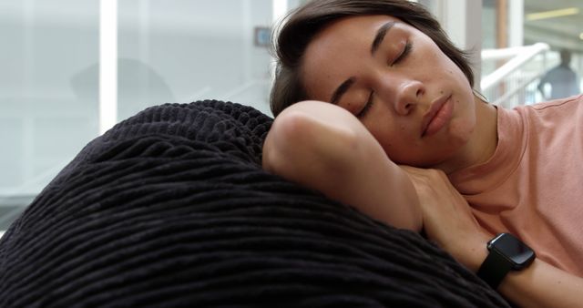 Woman is resting with head on comfortable black cushion, appearing peaceful and relaxed in an indoor setting. She wears a casual pink top and a smartwatch. Use for concepts of relaxation, comfort, and serenity, or for promoting rest-related products and environments.