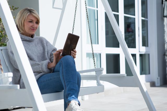 Woman sitting on a porch swing using a digital tablet on a sunny day. She is dressed casually in jeans and a sweater, enjoying leisure time outdoors. Ideal for use in lifestyle blogs, technology advertisements, and relaxation-themed promotions.