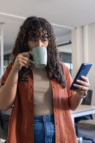 Curly-haired woman sipping coffee while checking smartphone in modern coworking office