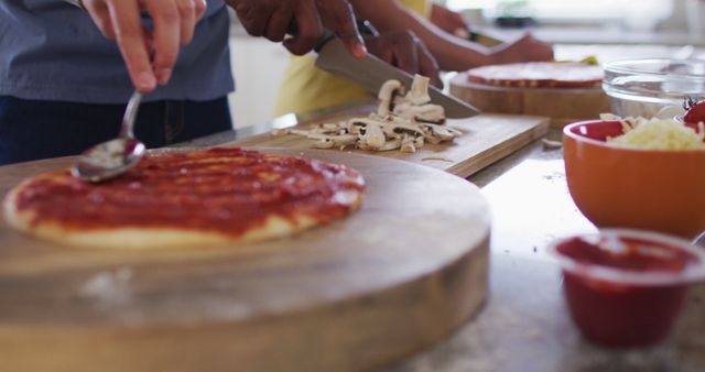 Hands seen chopping mushrooms and spreading tomato sauce on pizza dough. Multiple hands indicate a collaborative cooking activity in a domestic kitchen. Ideal for articles or advertisements related to family bonds, home cooking, recipes, or culinary experiences.