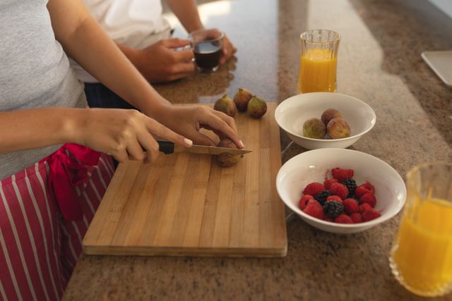 Couple slicing figs and preparing a healthy breakfast in their kitchen. Fresh fruit and juice are on the counter. Ideal for use in articles or advertisements about healthy living, home cooking, morning routines, or life during quarantine and social distancing.