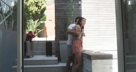 Young couple embracing on sunlit terrace with luggage, pink sun hat, planter and steps