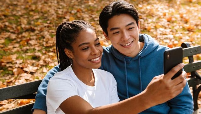 Happy black woman and asian man taking selfie on park bench among autumn leaves