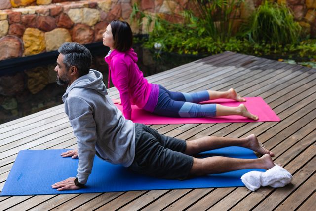 Diverse couple practicing yoga on a wooden deck in a garden. They are stretching and focusing on their fitness and well-being. This image can be used for promoting healthy lifestyles, fitness routines, yoga classes, outdoor activities, and wellness programs. It is also suitable for content related to spending quality time together at home.