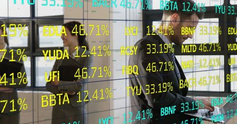Executive checking laptop amid live stock tickers with team collaborating behind glass