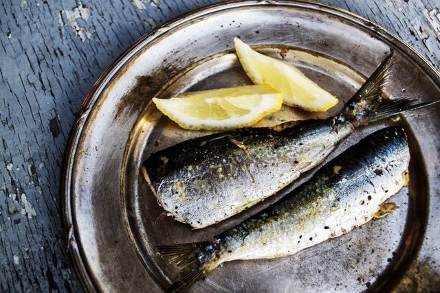 This image shows a close-up of two grilled sardines with two lemon slices on a rustic metal plate, placed on a weathered wooden surface. Ideal for use in food blogs, culinary magazines, seafood restaurant menus, or healthy eating promotions. Highlights the delicious and nutritious aspect of freshly prepared seafood.