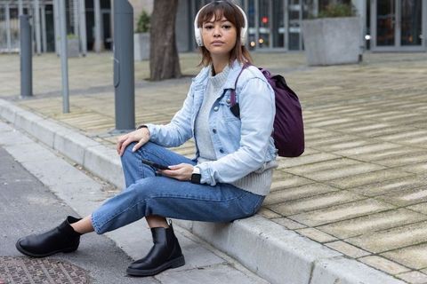 Young asian woman sitting on curb wearing headphones and denim jacket checking smartphone