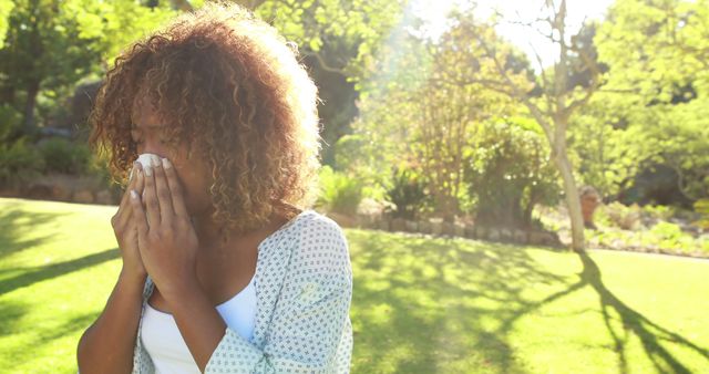 This photograph shows a woman sneezing while standing in a sunny park. She seems to be suffering from allergies. The vibrant green surroundings and sunlight make it ideal for themes related to nature, health, seasonal allergies, and wellness. The image can be used in blog posts, health articles, and advertisements related to allergy remedies or outdoor activities.