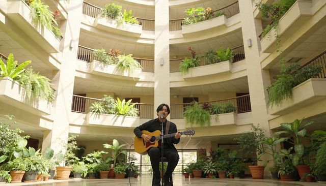 Playing asian musician performing acoustic guitar in sunlit atrium among indoor plants