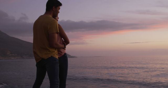 This image depicts a couple embracing while standing by the ocean during sunset. The warm hues of the evening sky, combined with the tranquil ocean background, create a serene and romantic atmosphere. This image is ideal for use in content related to romance, travel, scenic landscapes, or relaxation.