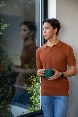 Asian man standing by glass window holding green mug, contemplative home moment