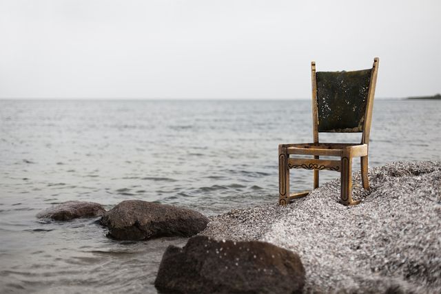 Vintage chair situated on a rocky beach next to the ocean shore. Ideal for use in decor blogs, antique furniture articles, or tranquil coastal scene contexts. This image conveys a sense of solitude, rustic charm, and peacefulness, suitable for illustrating themes of time passage, loneliness, or relaxation.