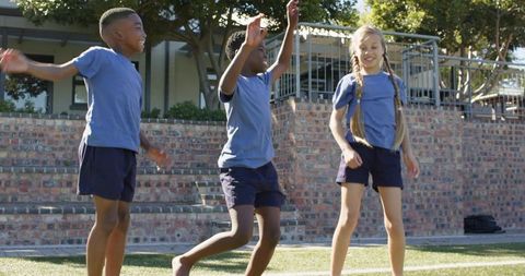Multiracial schoolchildren jumping on sunny sports field wearing light blue shirts and navy shorts