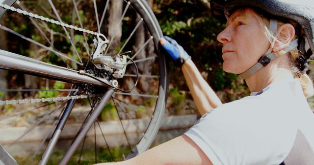 Senior woman wearing helmet repairing bicycle outdoors. Capturing active lifestyle and DIY spirit, this can be used in materials related to healthy living, senior activities, outdoor hobbies, and cycling maintenance.