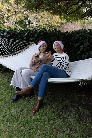 Friends wearing santa hats relaxing on white hammock in garden toasting holiday drinks