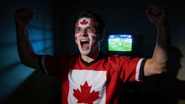 Canadian fan cheering and raising fists at home with face paint and maple leaf jersey