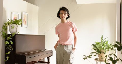 Young man standing beside upright piano in minimalist livingroom with plants, soft light
