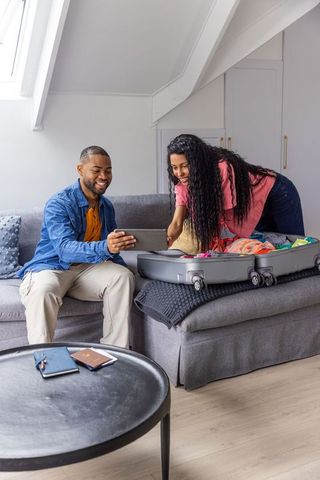 Young couple packing wheeled suitcase on gray sofa while checking tablet for trip