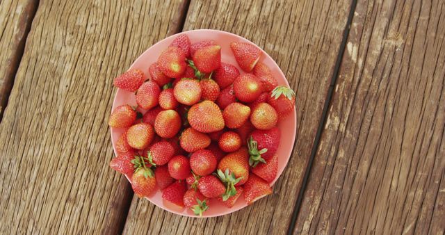 Bright red strawberries in a pink bowl set on a old wooden table. Ideal for use in food blogs, healthy living articles, advertisements for fresh produce, or recipes. Conveys themes of freshness, health, and summertime produce.