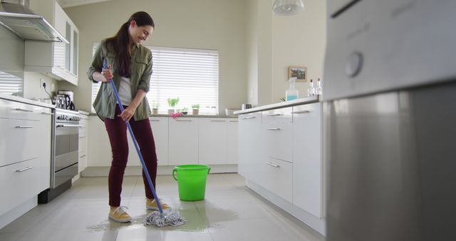 A woman is mopping the floor in a bright, modern kitchen during the daytime. She is smiling, suggesting she enjoys the task or has a positive attitude towards housework. The bright light from the window adds a cheerful atmosphere. This image can be used to represent house cleaning, domestic chores, home maintenance, or lifestyle content that emphasizes cleanliness and positive attitudes towards household responsibilities.