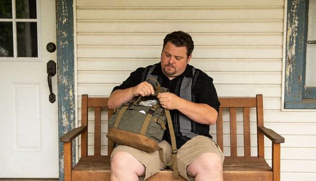 Man adjusting backpack straps on weathered porch bench preparing for trip