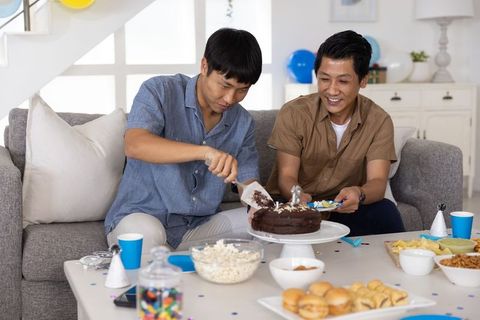 Two asian men cutting chocolate cake and serving snacks at casual home celebration