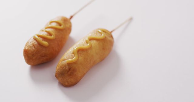 Golden brown corn dogs on white background with mustard drizzle. Ideal for food-related content, American recipes, fast food promotions, comfort food advertisements, and culinary blogs. Perfect for settings requiring a visual focus on casual dining and popular snacks.