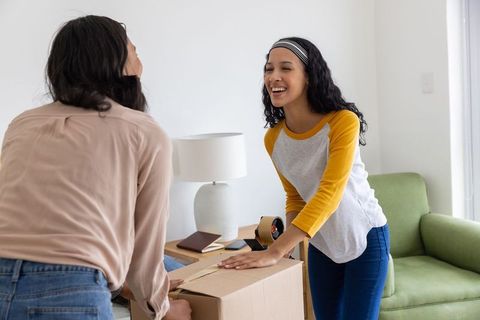 Diverse mother and daughter packing and sealing moving box, smiling in cozy living room