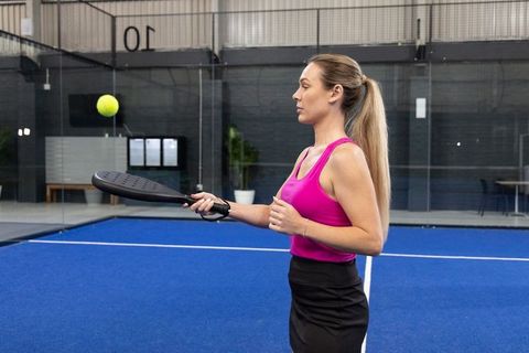 Woman practicing paddle tennis in pink top and black skirt on indoor blue court