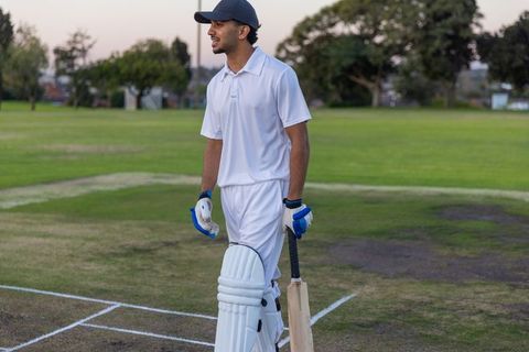 Indian cricketer standing on pitch at dusk holding bat wearing pads gloves and cap