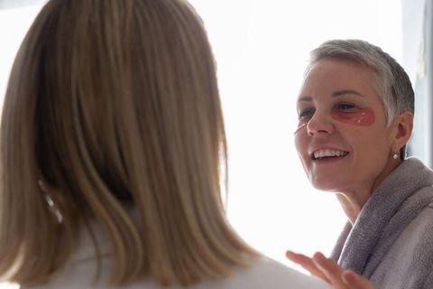 Mature woman wearing under-eye gel patches chatting with friend at vanity mirror