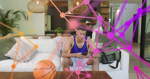 Teen male scrolling phone on sofa with basketball and duffel bag in modern living room